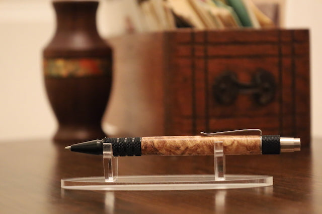 custom Pen with maple burl grip on a clear stand against a blurred backgroundl