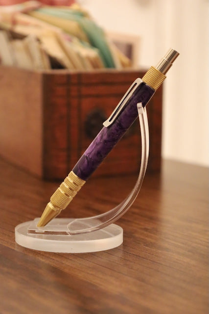 Purple and gold pen on a clear stand with a wooden box in the background. Custom pen
